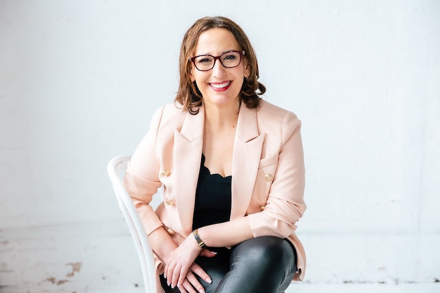Ms Calderone sitting on a chair in a white-walled studio smiling while wearing a peach blazer and black pants.