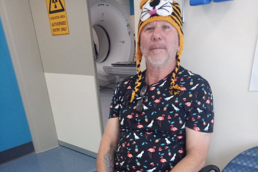 A man wears a knit tiger hat while sitting in a medical room next to a MRI machine and a mirror