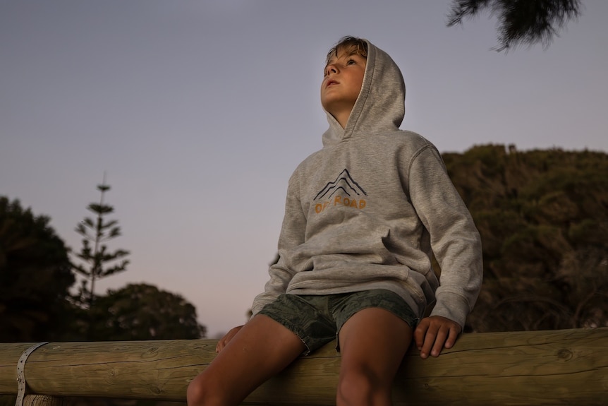 A young boy in a hoodie sits on a fence looking up into the night sky.