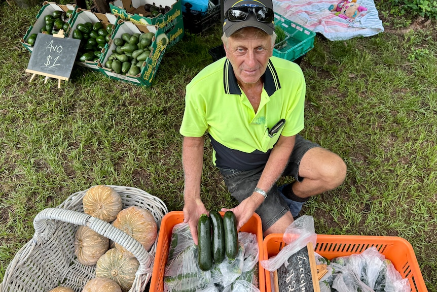 A man crouches next to zucchini and pumpkins in front of a market stall.