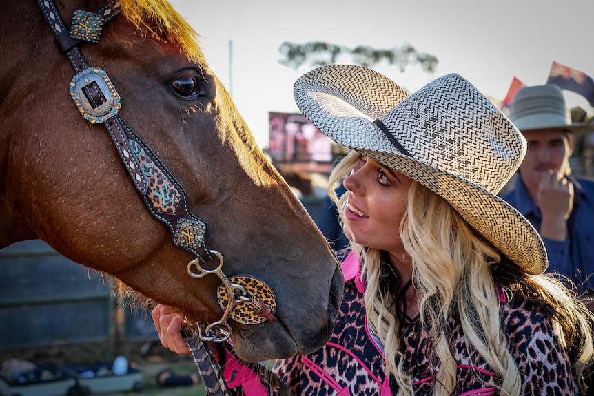 A woman wearing a cowboy hat looks fondly at a chestnut horse