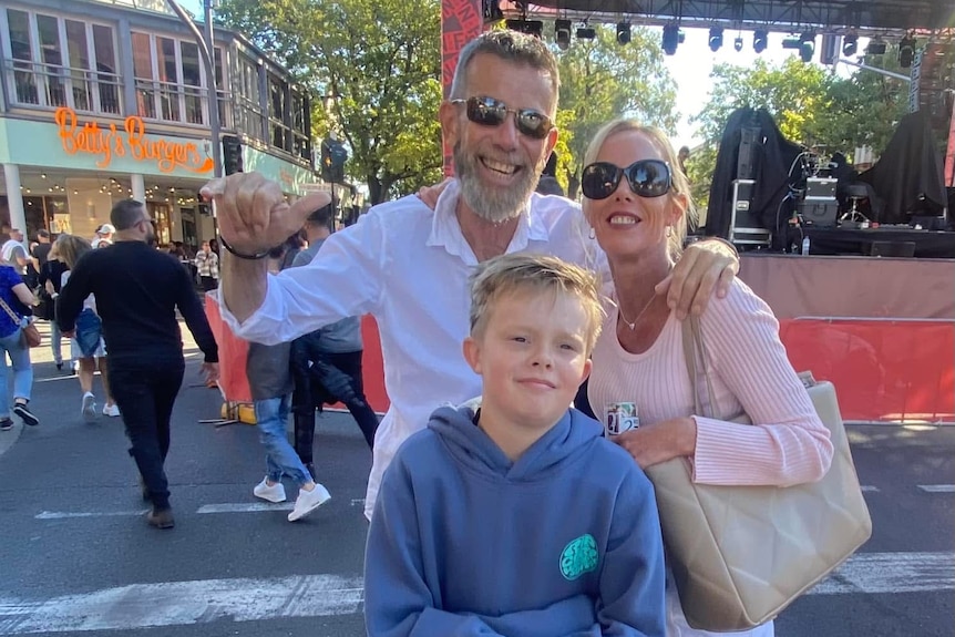 a man and a woman arms around each other behind a boy. They stand on a street in front of a makeshift stage