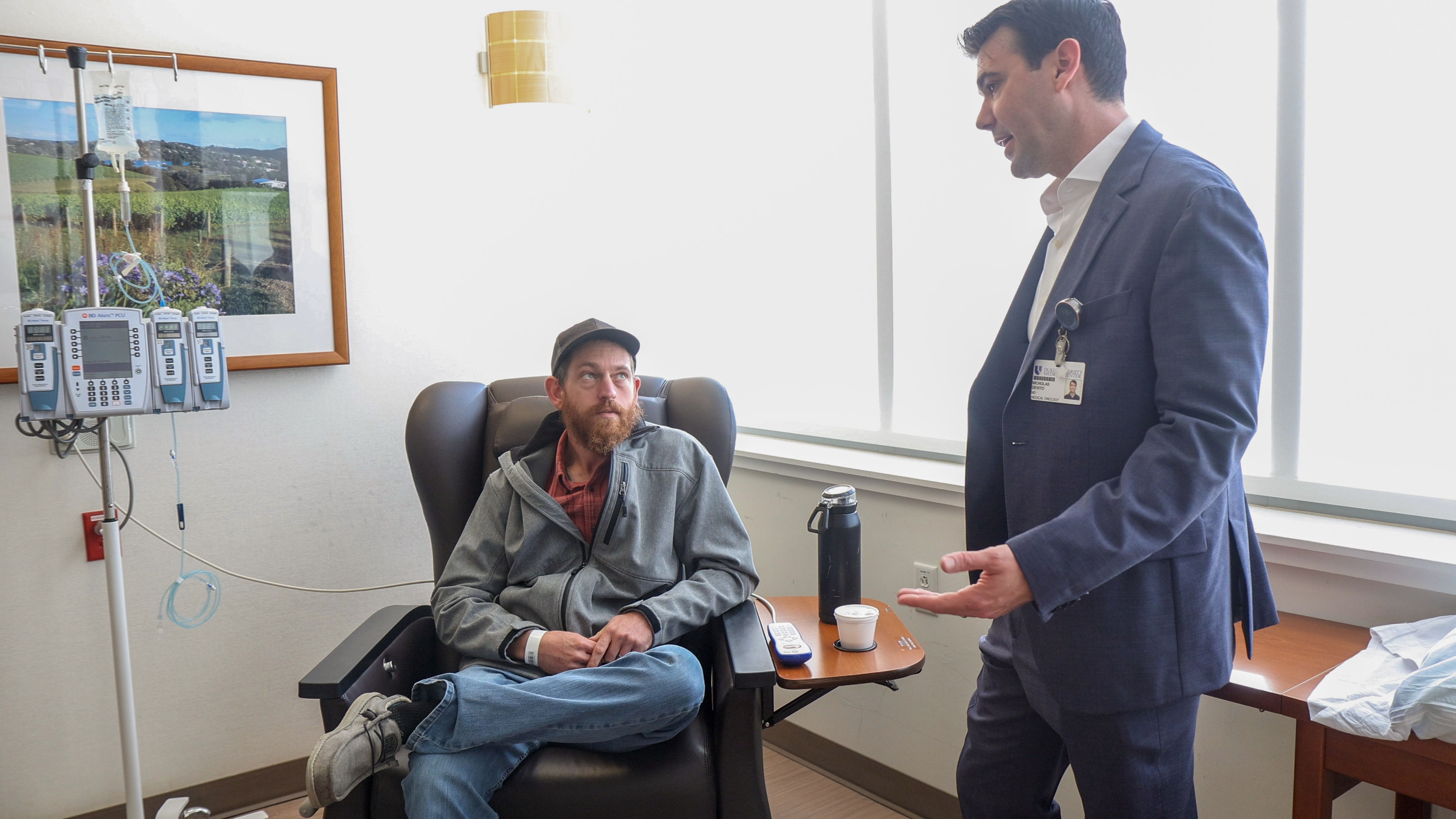 Patient seated in chair hooked up to IV while a doc in a suit stands talking to him