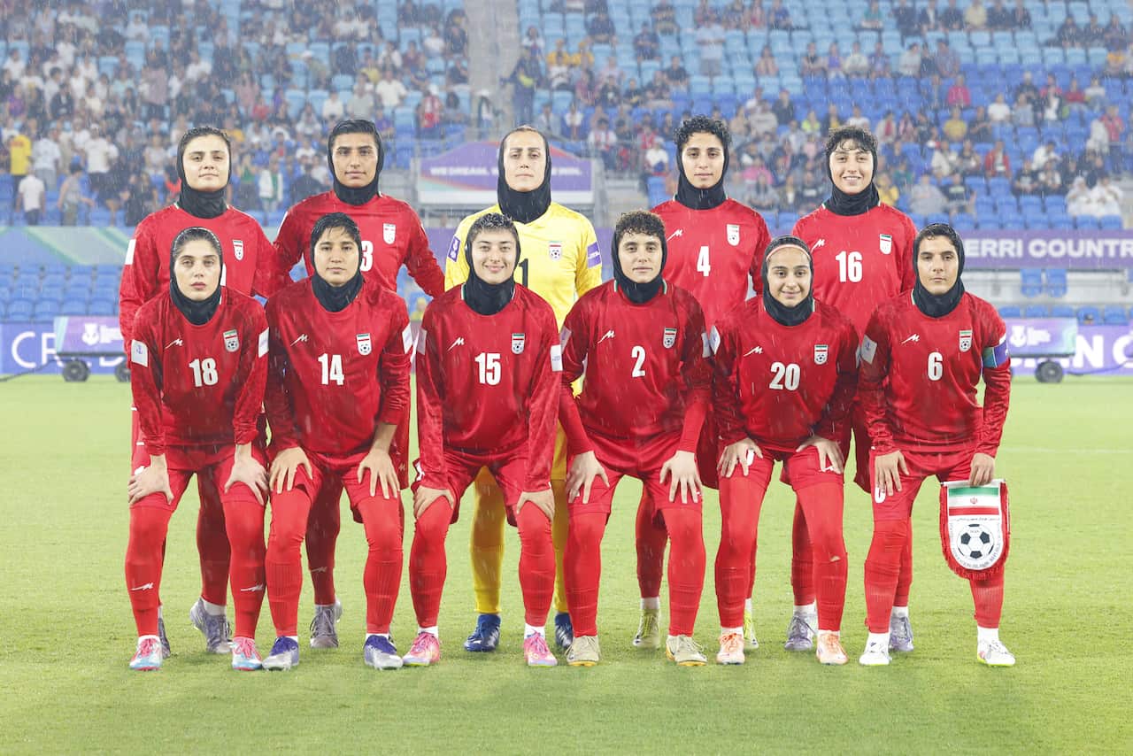 A team of female soccer players from the Iranian national team, wearing red kits and black hijabs, poses for a group photo on a rainy pitch before a match.
