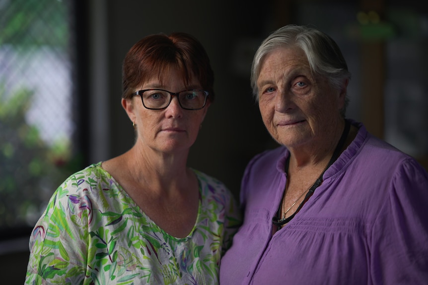 A middle-aged woman stands beside an older woman. Both look into camera with a serious facial expression.