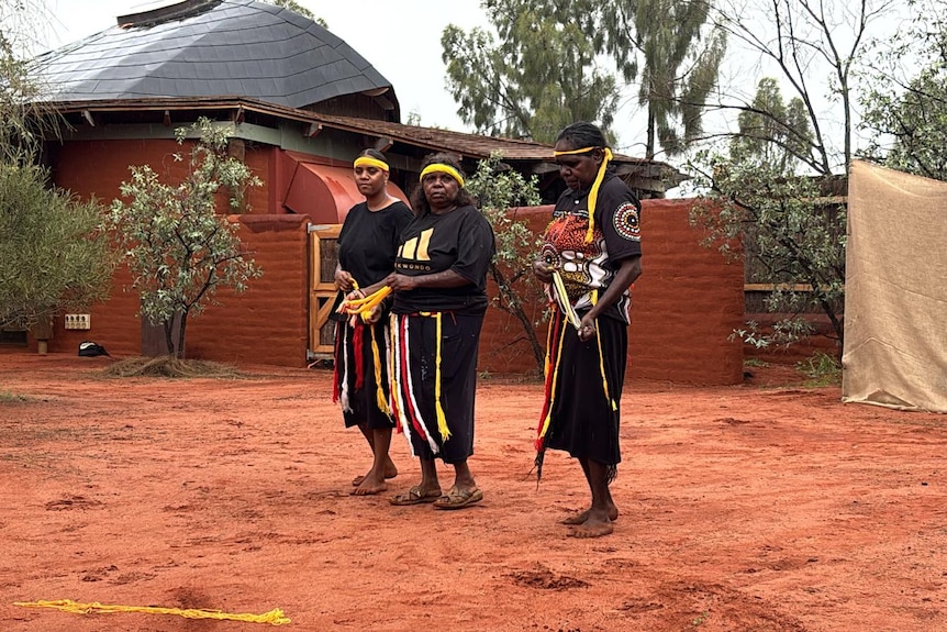 Three Indigenous women in headbands and matching, dark outfits stand and prepare to dance near a building in the outback.