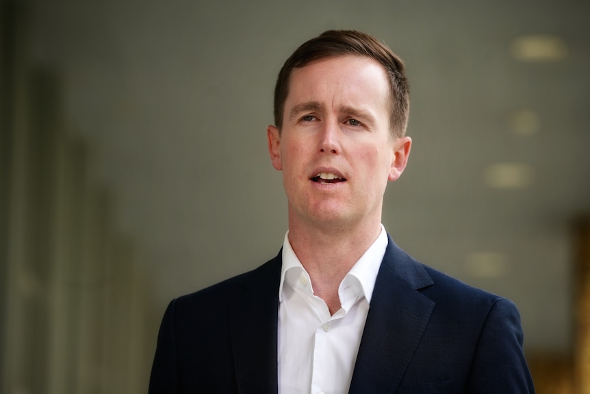 A man with short brown hair stands outdoors speaking seriously