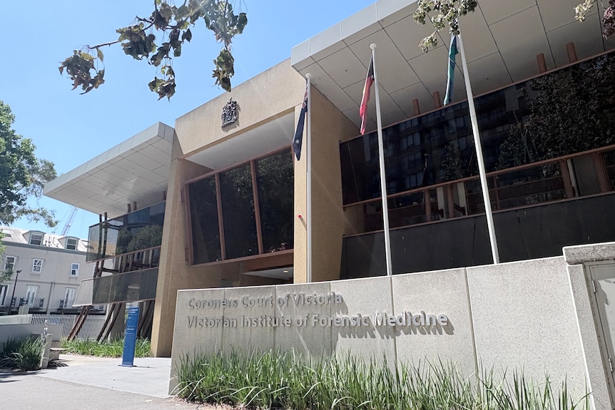 Flags hang limply from three poles near a building with a sign saying "Coroners Court of Victoria" on sunny day.