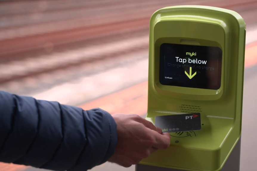 An arm in a blue jacket comes from left of frame and taps a myki card against a yellow card reader.