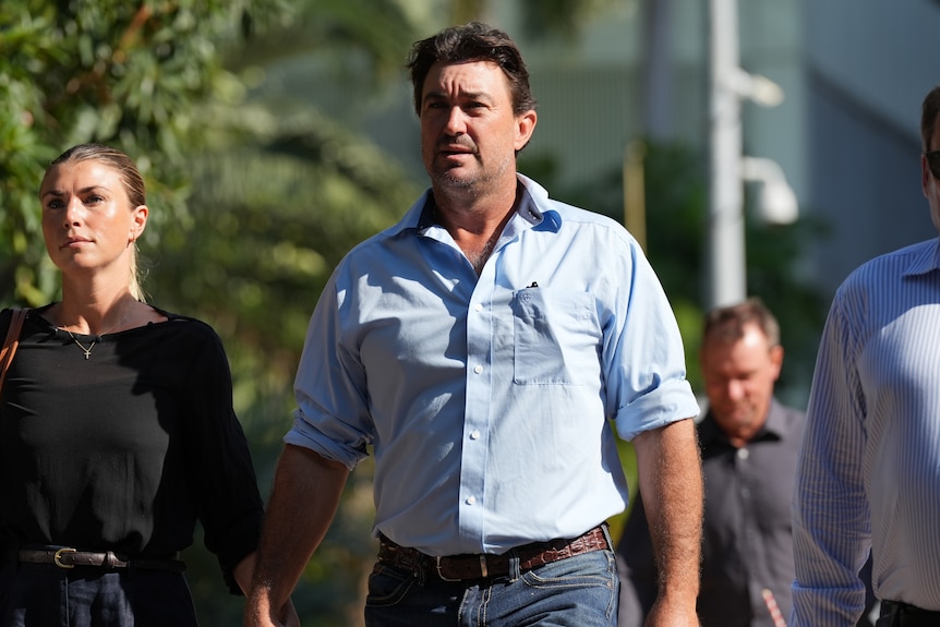 A man in a button-up shirt and jeans, walking towards the courthouse while holding his wife's hand.
