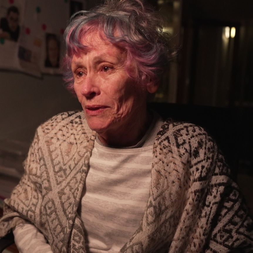 A mature woman with short hair and coloured fringe sits with a shall looking forward. She's seated, lit by singular light source