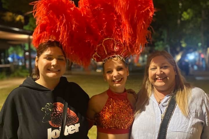 A young woman dressed in a red dance outfit with a large feather head piece smiles standing between two women.