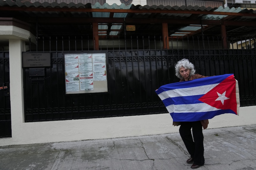 A woman with grey hair holding up a Cuban flag on a pathway next to a large black metal fence.