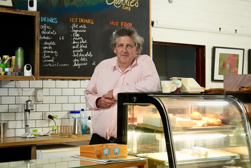 A man with grey hair wearing a pink shirt leans on a pastry fridge behind the counter of a regional general store.