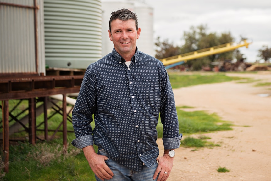 Man with white skin and blue shirt stands on rural property in front of water tank