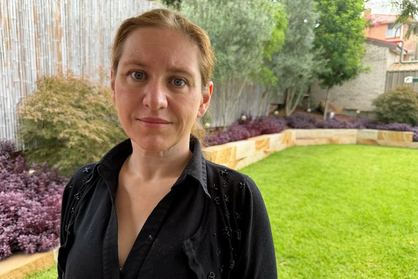 A woman in an open neck collared black shirt and hair pulled back looks at camera with a domestic garden behind