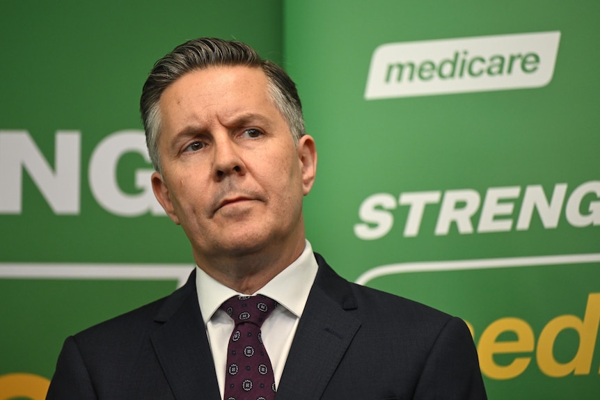 A politician in a suit and tie, standing in front of a green sign with the Medicare logo.