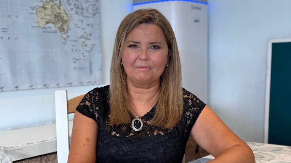 Dr Rae Duncan has long blonde/brown hair, a black top and a silver necklace with oval pendent. She is sat on a chair in a room with a global map on the wall.