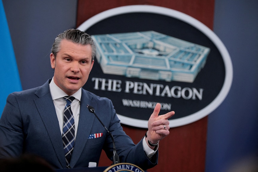 Pete Hegseth in a blue suit and tie with a US flag pocket square speaking with his hand in front of a Pentagon sign.