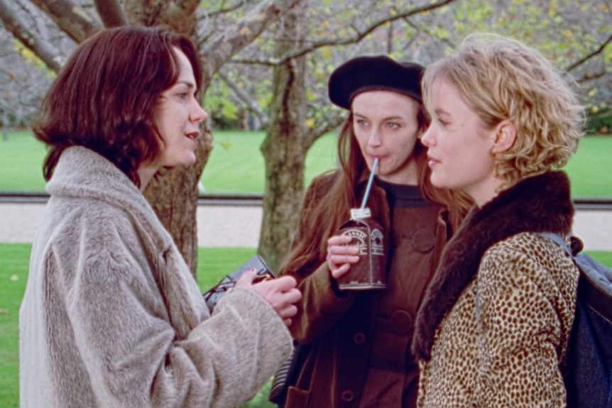 Film still of three women in 90s coats standing on a grassy lawn