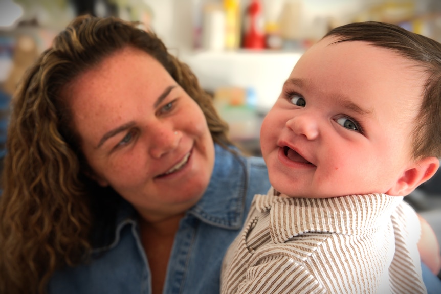A headshot of a baby in mum's arms. She's looking at him and the baby is smiling.