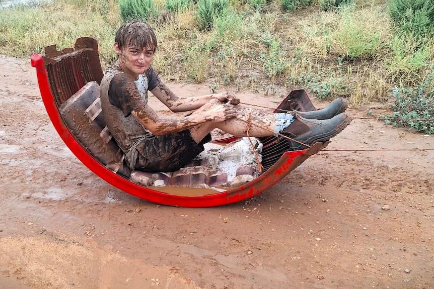 A mud-covered boy sits in a curved sled on a dirt track.