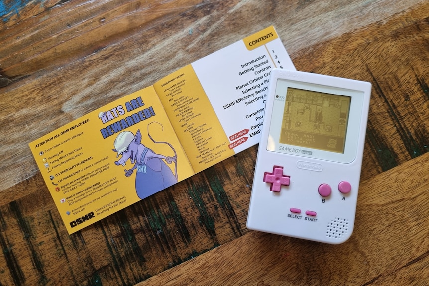 A game boy console sits on a table displaying a game on it's screen with an open game manual behind it. 