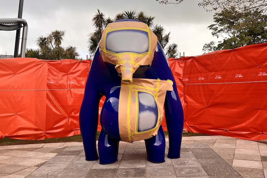 A large blue sculpture with gold tape and grey markings on it in front of an orange fence.