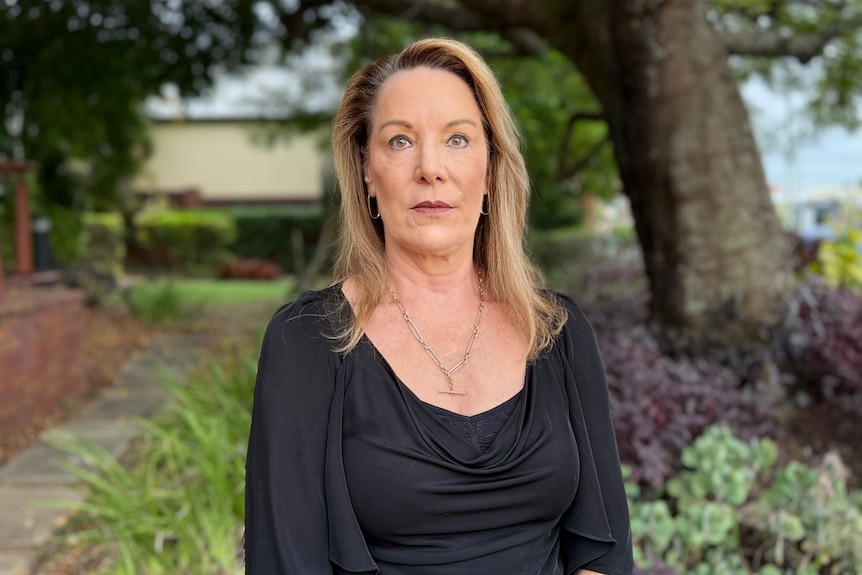 A woman standing in front of a leafy garden with a sombre expression