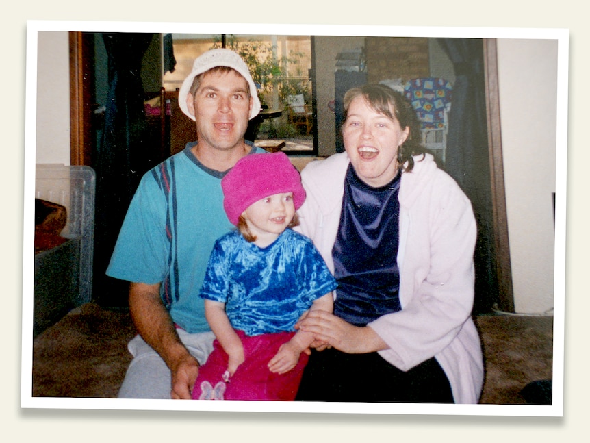 A man with a white hat on smiling, sitting next to a small girl with a pink beanie next to her mother in a white cardigan.
