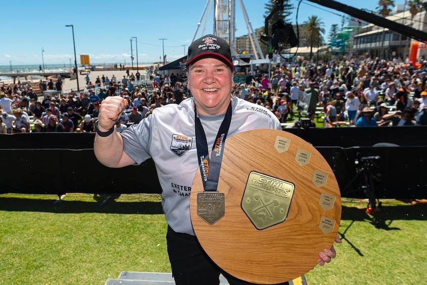 A woman wearing a grey t-shirt and black cap holds a wooden shield trophy and wears a medal around her neck.