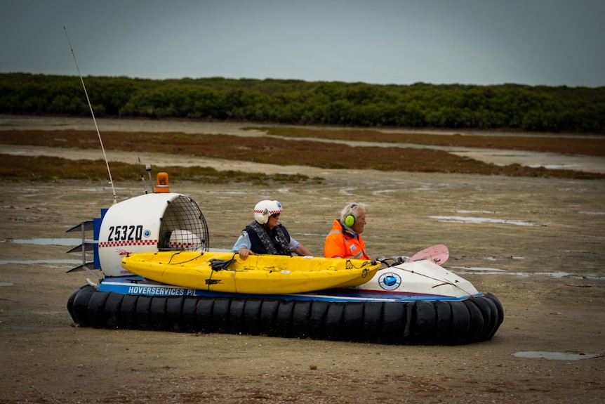 Two officers in helmet on a hovercraft, one of them holds a kayak attached to the side, on wetlands