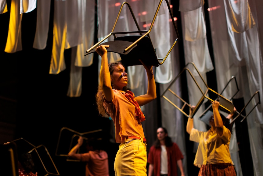 Young woman holds chair above head, other actors in background