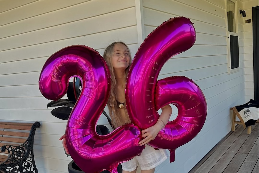 A young woman with long blond hair holds bright pink balloons in the shape of 26.