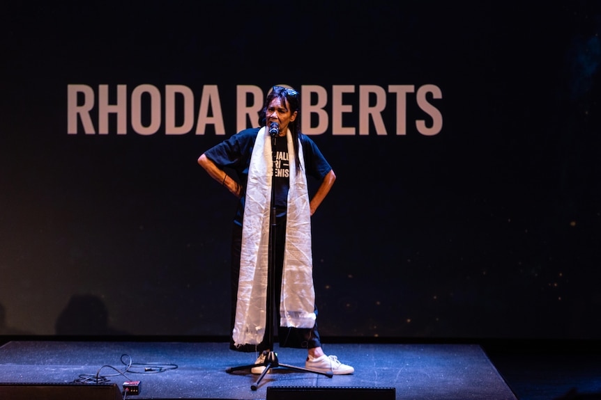 Rhoda Roberts AO on stage with hands on hips and her name written on the wall behind her