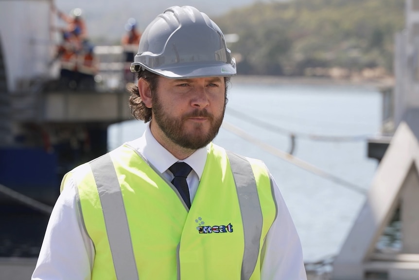 Felix Ellis stands at a wharf wearing a hard hat and a yellow Hi-Vis vest.