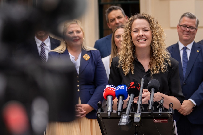 A woman speaks into a microphone with a camera in the foreground. Behind her is a group of people.