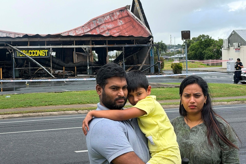 A man and a woman hold their young son in front of a burnt building, looking sad.