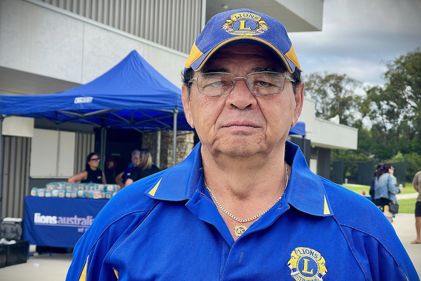 Man in a blue lions shirt and cap in front of barbecue stall.