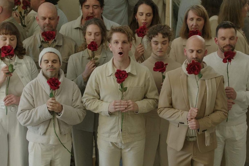 A group of singers dressed in white and beige face the camera holding large red flowers. Most have closed eyes as they sing.