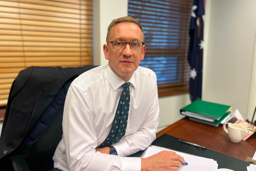 A white man with short brown hair and glasses sitting in an office. He's wearing a shirt and tie