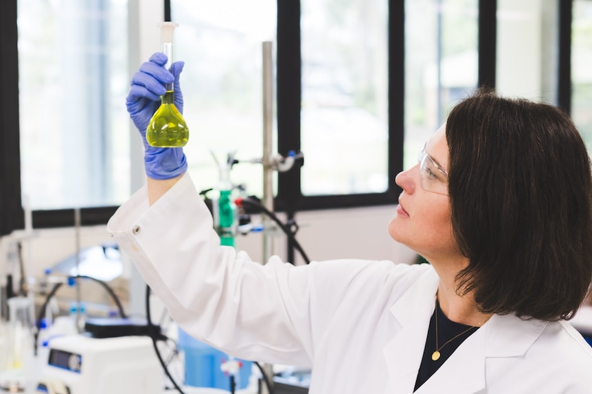 A researcher in a lab peers at a beaker of green liquid. 
