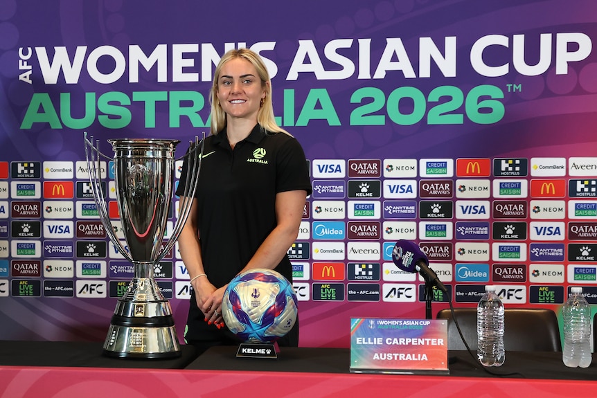 Matildas vice captain Ellie Carpenter smiles and poses behind the Women's Asian Cup trophy and a ball