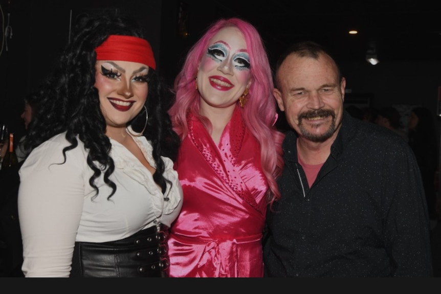 A man stands with two drag queens smiling at the camera.