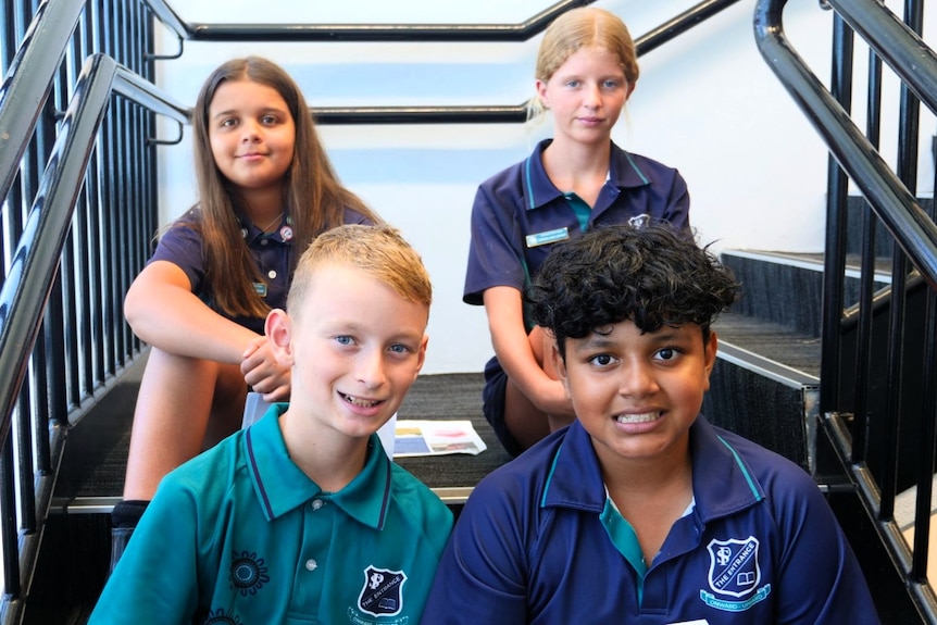 Two boys in blue and green school uniforms with two girls sitting behind them on a stair case.