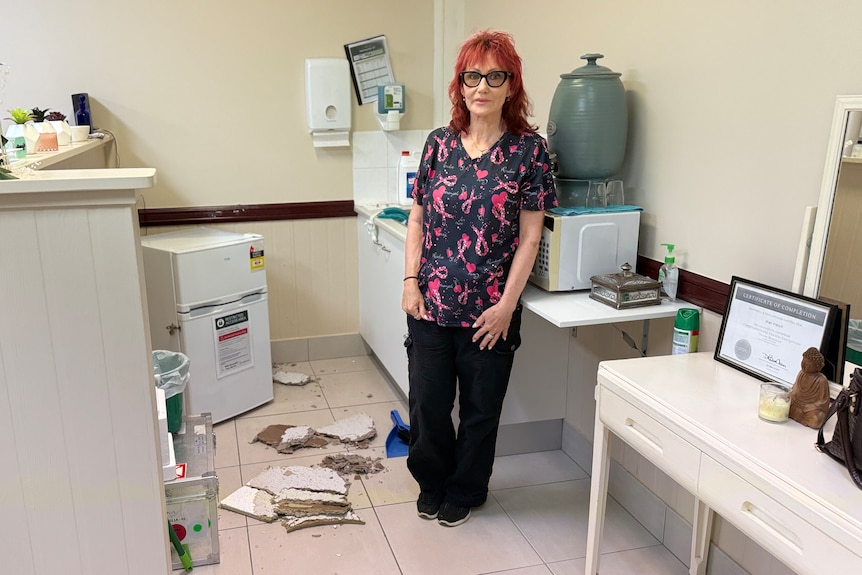 A red-haired woman in glasses stands next to some fallen tiles on a kitchen floor.