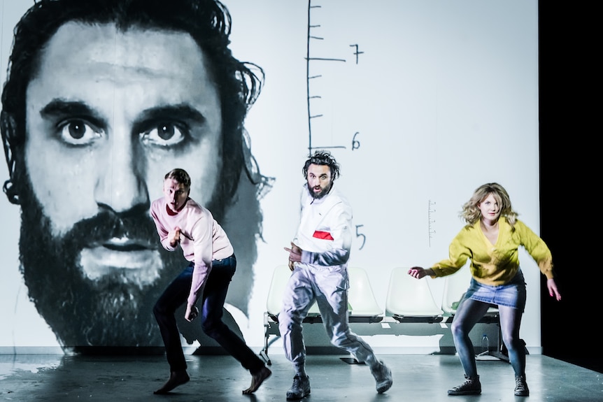 On stage, three people, including a Brazilian actor, move as if running, a portrait of the Brazilian man on the screen behind.