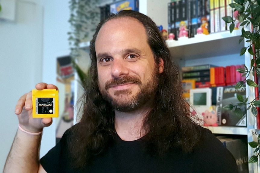 A man smiles towards the camera holding a yellow game boy cartridge.