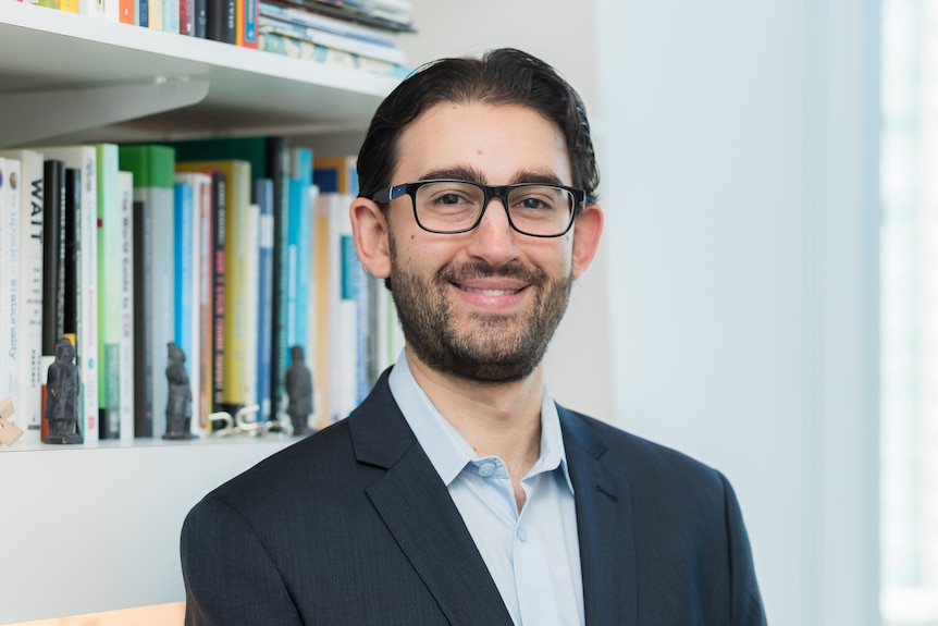 A man wearing glasses and a suit looks at the camera and smiles