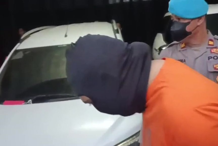 A man in a balaclava stoops his head as a police officer escorts him past a car.